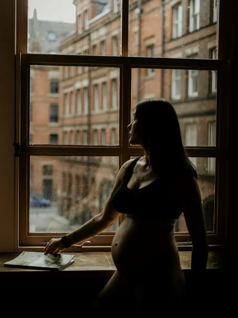 Eine schwangere Frau steht im Gegenlicht an einem Fenster